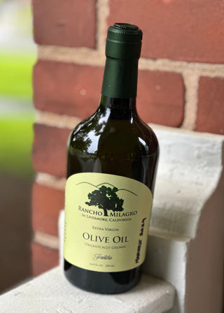Bottle of Rancho Milagro Frantoio olive oil with a label on a ledge against a brick wall.