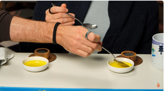 Two hands dipping metal spoons into two small white saucers with bright yellow olive oil samples in them. 