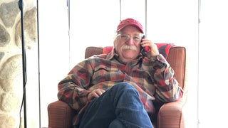 Smiling man wearing a long-sleeved plaid flannel shirt and jeans, with silver walrus mustache, silver hair visible under his red ball cap with Sundance Film Festival logo, seated on a well-cushioned chair, phone to his ear.