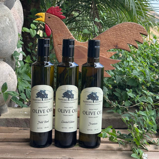 Three dark glass bottles of Rancho Milagro Extra Virgin Olive Oil arranged on a wooden surface outdoors, with green foliage, a wooden rooster decoration, and stone garden elements in the background.