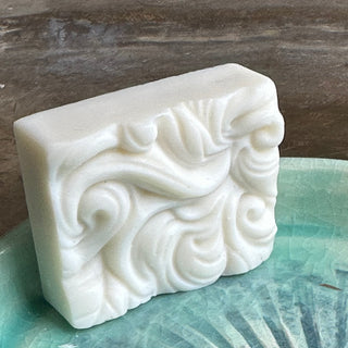 Rectangular soap with swirlly, wavy relief on one face, resting on its side in a green ceramic dish.