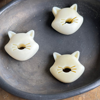 3 small soaps, each identical: cat's wide-cheek head and pointed ears, with center cut out for nose+mouth and whiskers, resting on a black ceramic dish.