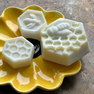 Three hexagonal soaps, two small and one large. The small ones are a bee (top) and honeycomb (bottom). At right is the large honeycomb, with a bee atop. All are resting on a shiny yellow scalloped dish.
