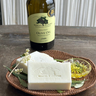 2 bars of soap, olive oil in a glass saucer. Olive blossoms and leaves scattered around the handwoven platter. Rancho Milagro olive oil bottle in the background.