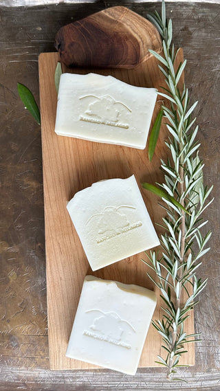 Three bars of soap resting on a wooden tray, fresh rosemary branch beside.