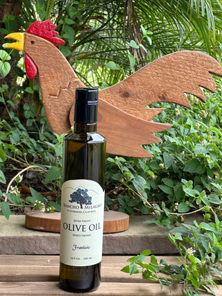 A bottle of Frantoio extra virgin olive oil from Rancho Milagro stands on a wooden surface in front of a decorative wooden rooster cutout, with lush green garden foliage in the background.