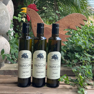 Three dark glass bottles of Rancho Milagro Extra Virgin Olive Oil arranged on a wooden surface outdoors, with green foliage, a wooden rooster decoration, and stone garden elements in the background.