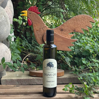 A bottle of Rancho Milagro Extra Virgin Olive Oil stands on a wooden surface in front of a decorative wooden rooster sculpture, surrounded by green foliage and garden stones.