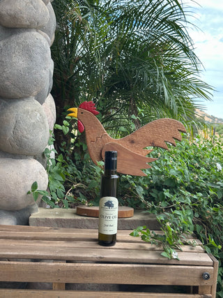A 250ml bottle of olive oil stands on a wooden crate outdoors, with a carved wooden rooster decoration behind it and green plants and a stone column in the background.
