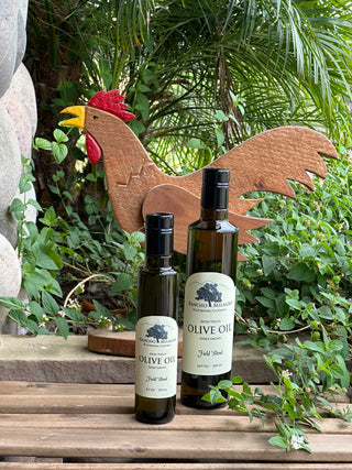 Two dark glass bottles of olive oil, smaller on left and larger on the right, with pale yellow labels and black caps, displayed in front of a wooden rooster stand outdoors against a background of green foliage.