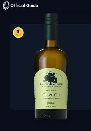 A 500ml (older) bottle of Rancho Milagro Extra Virgin Olive Oil centered on a dark navy background with an 'Official Guide' banner for the New York International Olive Oil Competition at the top and a gold 2026 award badge on the left.