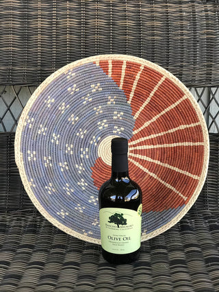 Dark bottle of extra virgin olive oil on a wicker bench with a hand woven stars-and-stripes-theme basket behind the bottle. 